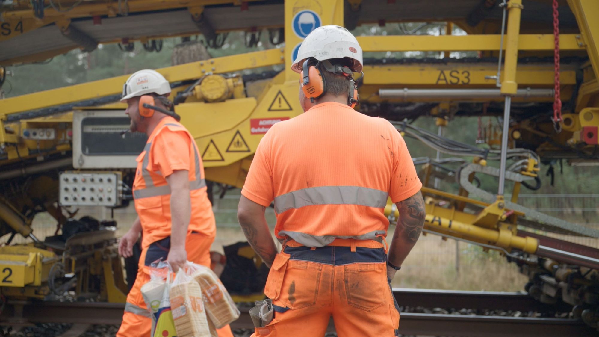 Twee monteurs in oranje werkkleding naast een gele spoorvernieuwingsmachine